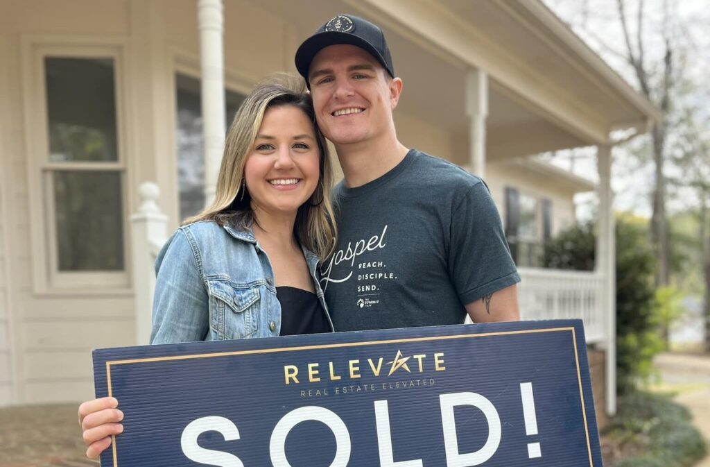 A young couple holds a "Sold" sign after working and selling their home with Relevate