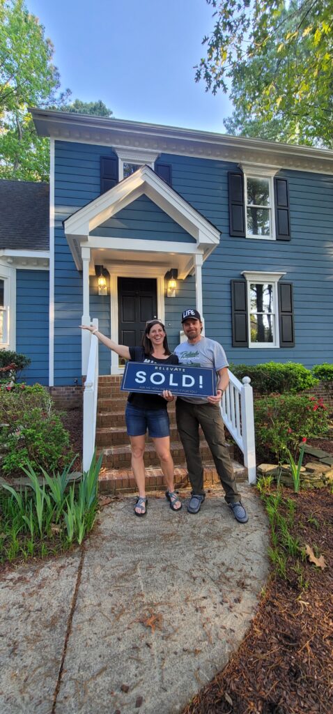 two satisfied home buyers in front of blue house holding a sold sign