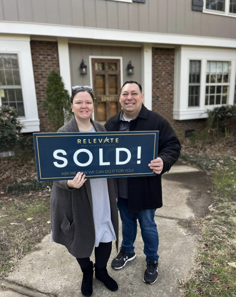 Two satisfied home buyers hold sold sign in front of home